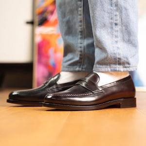 May include: A pair of black leather penny loafers. The shoes are shiny and have a classic design. The image shows the shoes from the side, with the top of the shoes visible. The shoes are worn with light blue jeans and white socks.