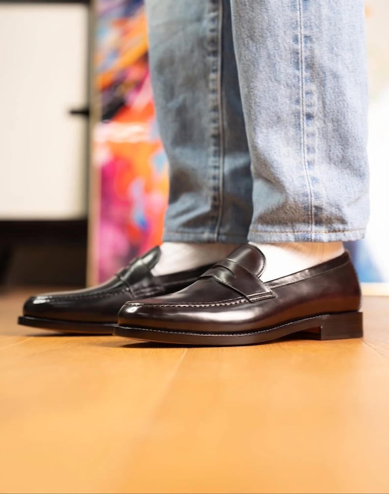 May include: A pair of black leather penny loafers. The shoes are shiny and have a classic design. The image shows the shoes from the side, with the top of the shoes visible. The shoes are worn with light blue jeans and white socks.