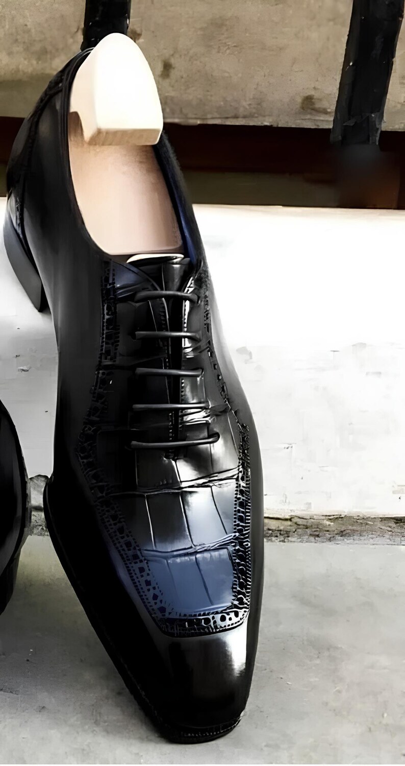 May include: A close-up of a black leather dress shoe with a glossy finish. The shoe features a textured, crocodile-like pattern on the toe and heel, with a classic lace-up design. A wooden shoe tree is inserted inside the shoe.