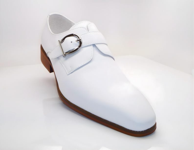 May include: A white leather shoe with a buckle closure. The shoe has a brown sole and heel. The buckle is silver-colored. The shoe is a formal style.