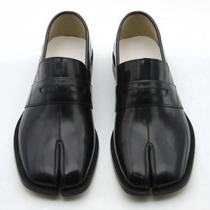 May include: Pair of black leather split-toe loafers. The shoes have a glossy finish, a strap across the top, and a white interior. The soles are black and the shoes are displayed on a white background.