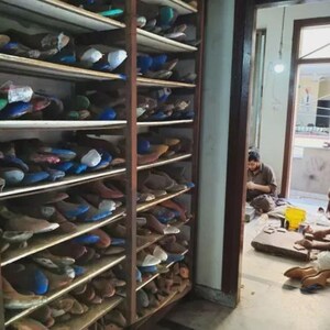 May include: Wooden shelves filled with shoe lasts in various colors and shapes. The shelves are part of a workshop, with two people visible in the background working on footwear. The lasts are used to shape shoes during the manufacturing process.