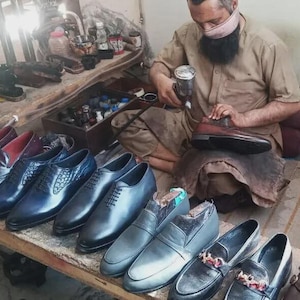 Peut inclure: Une collection de chaussures habill&eacute;es en cuir dans diverses nuances de noir, marron et bleu, comprenant des richelieus &agrave; lacets, des mocassins et des mod&egrave;les &agrave; enfiler dispos&eacute;s sur un &eacute;tabli en bois pr&egrave;s d'une personne utilisant un pistolet.