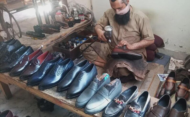Peut inclure: Une collection de chaussures habill&eacute;es en cuir dans diverses nuances de noir, marron et bleu, comprenant des richelieus &agrave; lacets, des mocassins et des mod&egrave;les &agrave; enfiler dispos&eacute;s sur un &eacute;tabli en bois pr&egrave;s d'une personne utilisant un pistolet.
