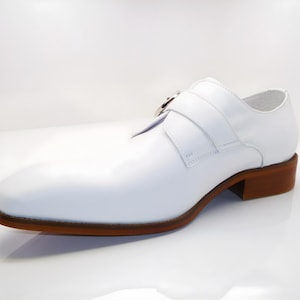 May include: A white leather shoe with a brown sole and heel. The shoe features a strap with a silver buckle across the top. The design is a classic style, suitable for formal occasions.