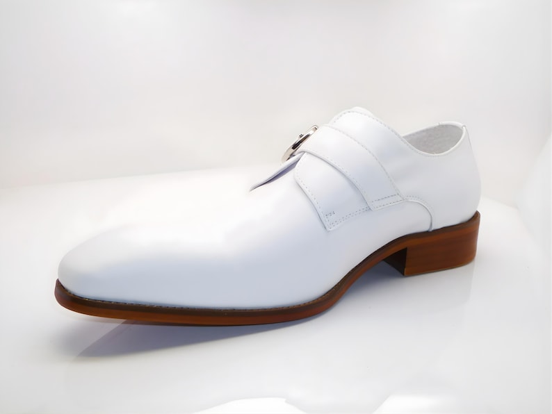 May include: A white leather shoe with a brown sole and heel. The shoe features a strap with a silver buckle across the top. The design is a classic style, suitable for formal occasions.