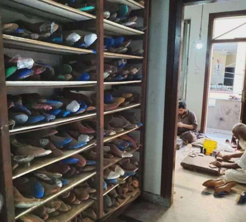 May include: A wooden shelving unit filled with shoe lasts in various colors. The lasts are arranged on multiple shelves, with a glimpse of a workshop in the background where shoemakers are working on footwear.