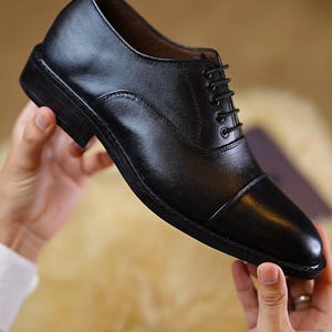Peut inclure: Une chaussure oxford en cuir noir avec un bout droit, un petit talon empil&eacute; et des lacets noirs fins passant par cinq paires d'&oelig;illets m&eacute;talliques. La chaussure pr&eacute;sente une finition lisse et polie ainsi qu'une silhouette structur&eacute;e.