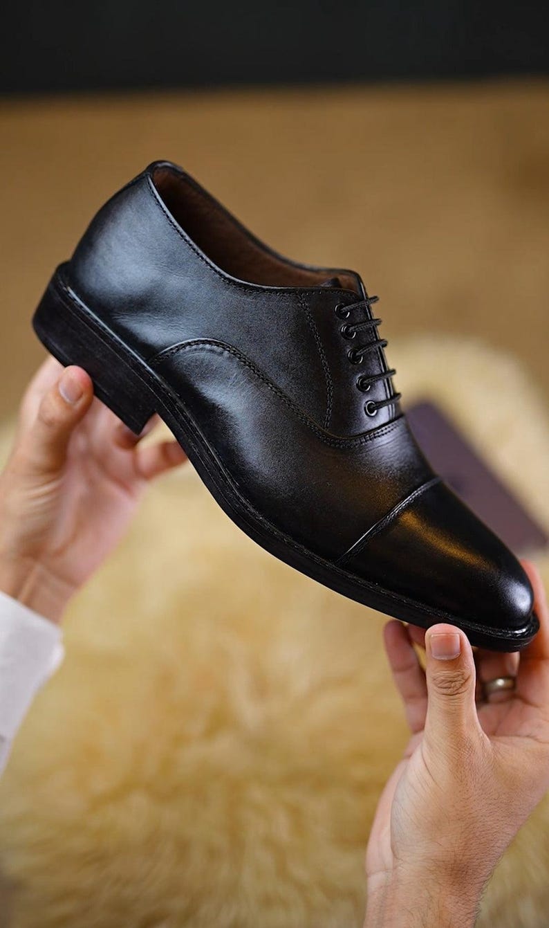 Peut inclure: Une chaussure oxford en cuir noir avec un bout droit, un petit talon empil&eacute; et des lacets noirs fins passant par cinq paires d'&oelig;illets m&eacute;talliques. La chaussure pr&eacute;sente une finition lisse et polie ainsi qu'une silhouette structur&eacute;e.