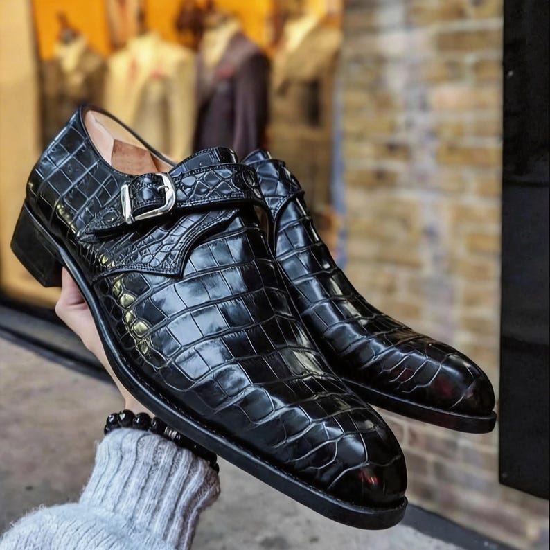 May include: A pair of black leather dress shoes with a crocodile skin texture. The shoes feature a buckle closure and a low heel. The glossy finish reflects the light, highlighting the intricate pattern of the leather.
