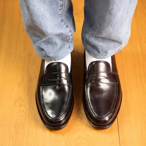May include: A pair of black leather penny loafers with a glossy finish. The shoes are worn with light blue denim jeans and white socks. The loafers are on a wooden floor.