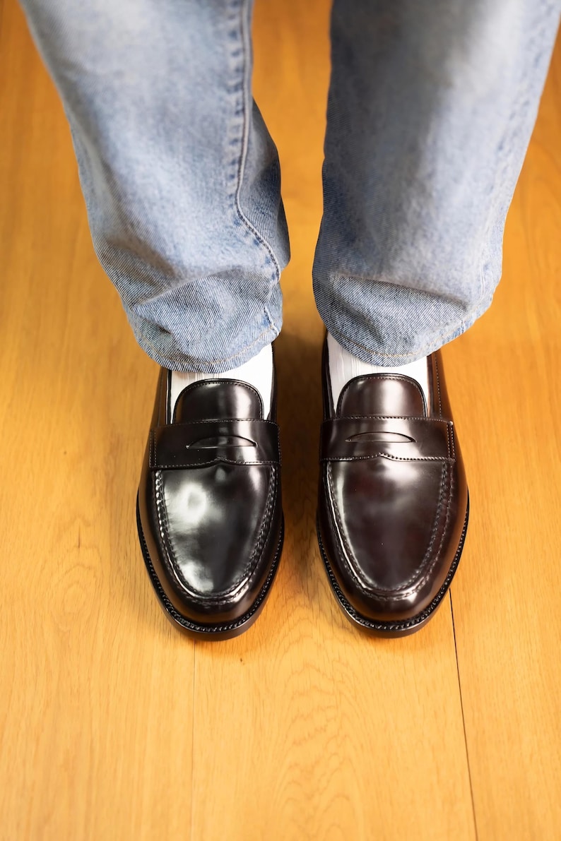May include: A pair of black leather penny loafers with a glossy finish. The shoes are worn with light blue denim jeans and white socks. The loafers are on a wooden floor.