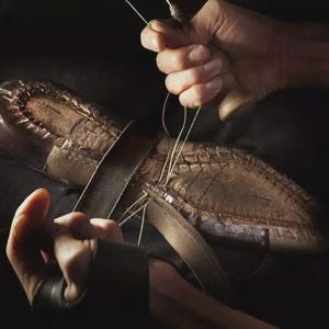 Peut inclure: Une semelle de chaussure en cuir marron fonc&eacute; cousue &agrave; la main avec du fil clair. Le processus implique un manche d'al&ecirc;ne en bois et une lani&egrave;re de cuir positionn&eacute;e sur la semelle, montrant la construction manuelle complexe de la chaussure.