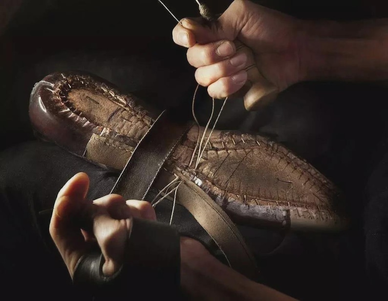 Peut inclure: Une semelle de chaussure en cuir marron fonc&eacute; cousue &agrave; la main avec du fil clair. Le processus implique un manche d'al&ecirc;ne en bois et une lani&egrave;re de cuir positionn&eacute;e sur la semelle, montrant la construction manuelle complexe de la chaussure.