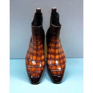 May include: A pair of ankle boots featuring a glossy, gradient brown finish with a textured crocodile skin pattern. These boots have a pointed toe, a pull tab at the back of the collar, and a dark sole with visible stitching along the edge.