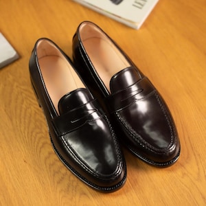 May include: A pair of black leather penny loafers with a glossy finish. The shoes are displayed on a wooden surface, with a magazine in the background. The loafers have a classic design with a strap across the vamp.