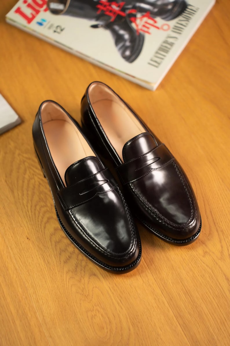 May include: A pair of black leather penny loafers with a glossy finish. The shoes are displayed on a wooden surface, with a magazine in the background. The loafers have a classic design with a strap across the vamp.