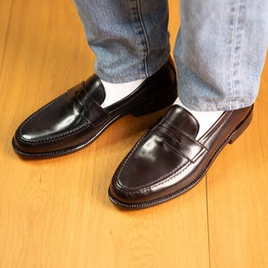 May include: A pair of black leather penny loafers with a glossy finish. The shoes have a classic design with a strap across the vamp and are worn with white socks and blue jeans. The shoes are on a wooden floor.
