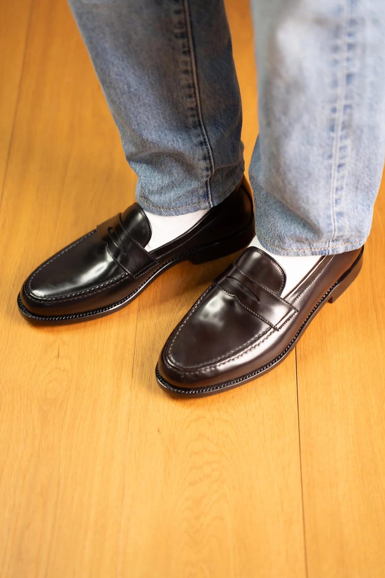 May include: A pair of black leather penny loafers with a glossy finish. The shoes have a classic design with a strap across the vamp and are worn with white socks and blue jeans. The shoes are on a wooden floor.