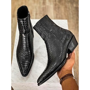 May include: A pair of black ankle boots featuring a textured snakeskin pattern. The boots have a pointed toe, a side zip closure, and a stacked heel. The material has a glossy finish, and the design includes a slim silhouette with a high shaft.