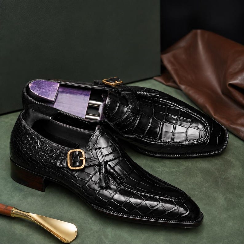 K&ouml;nnte beinhalten: Ein Paar schwarze Lederschuhe mit Krokodilmuster. Die Schuhe haben einen Schnallenverschluss mit goldfarbener Schnalle und Quastendetail. Die Schuhe stehen auf einer gr&uuml;nen Oberfl&auml;che.
