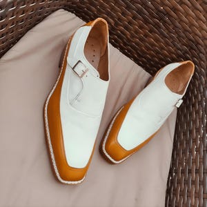 May include: A pair of white and tan leather shoes with a buckle fastening. The shoes have a square toe and a tan border. The shoes are on a beige surface.