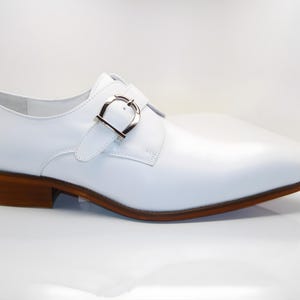 May include: A white leather shoe with a silver buckle closure. The shoe has a brown sole and heel. The design is a classic dress shoe style, suitable for formal occasions. The shoe's heel measures approximately 1 inch.