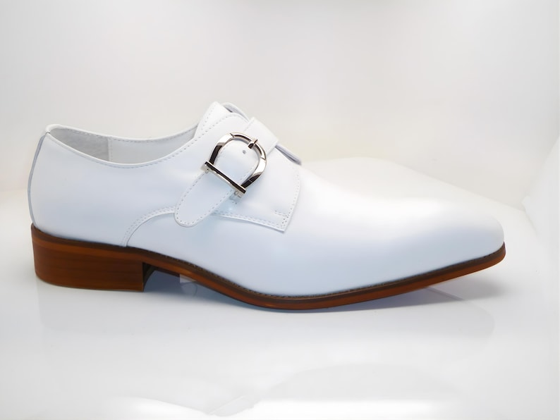 May include: A white leather shoe with a silver buckle closure. The shoe has a brown sole and heel. The design is a classic dress shoe style, suitable for formal occasions. The shoe's heel measures approximately 1 inch.