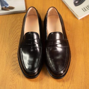 May include: A pair of black leather penny loafers with a glossy finish. The shoes are displayed on a wooden surface, with a magazine visible in the background. The loafers have a classic design, suitable for formal or casual wear.