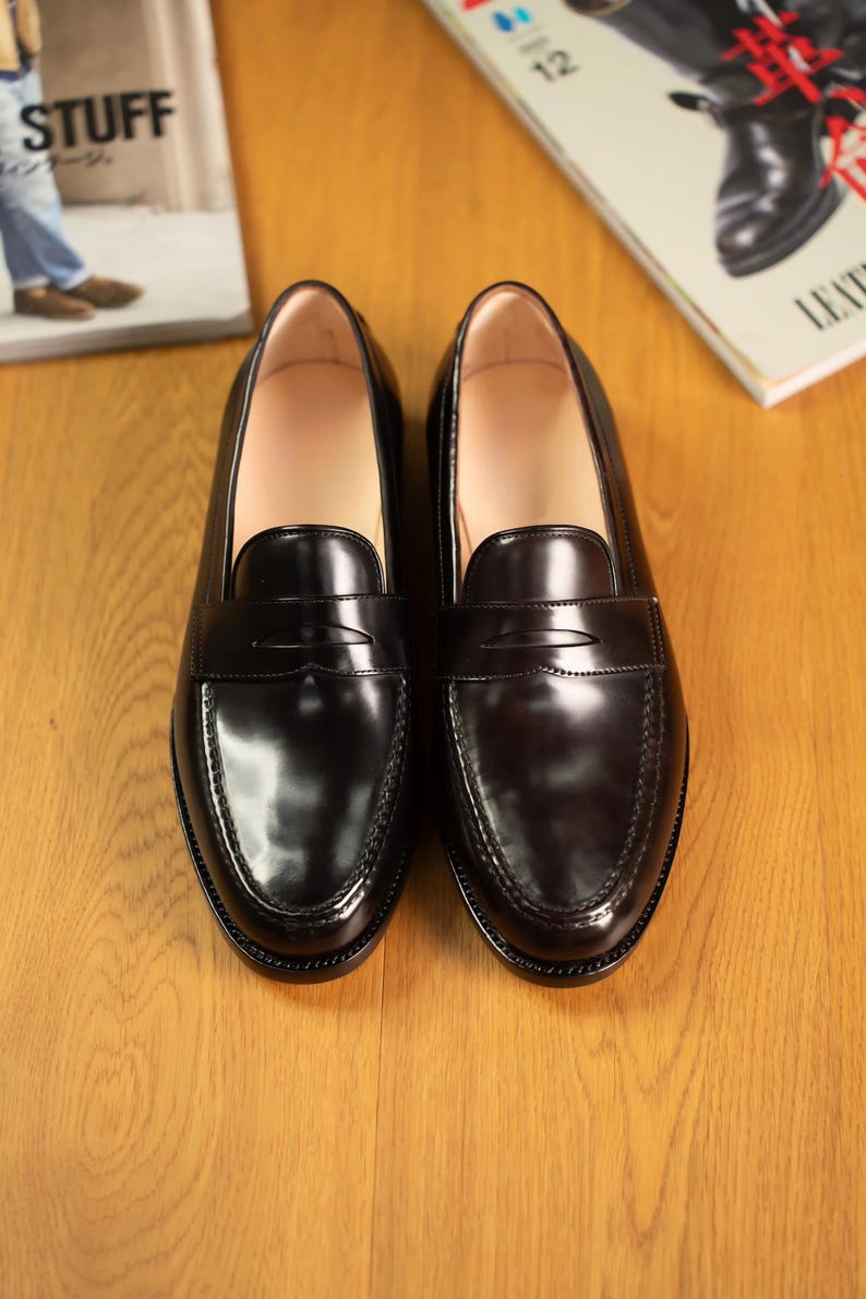 May include: A pair of black leather penny loafers with a glossy finish. The shoes are displayed on a wooden surface, with a magazine visible in the background. The loafers have a classic design, suitable for formal or casual wear.