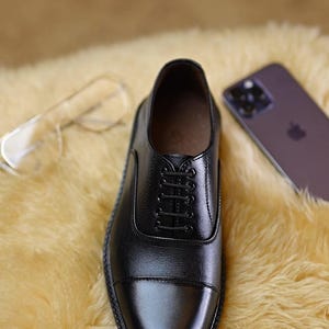 Peut inclure: Une chaussure richelieu en cuir noir avec un bout droit, dot&eacute;e d'un syst&egrave;me de la&ccedil;age ferm&eacute; avec cinq paires d'&oelig;illets et de fins lacets noirs. La chaussure pr&eacute;sente une finition lisse et polie ainsi qu'une tr&eacute;pointe cousue visible.