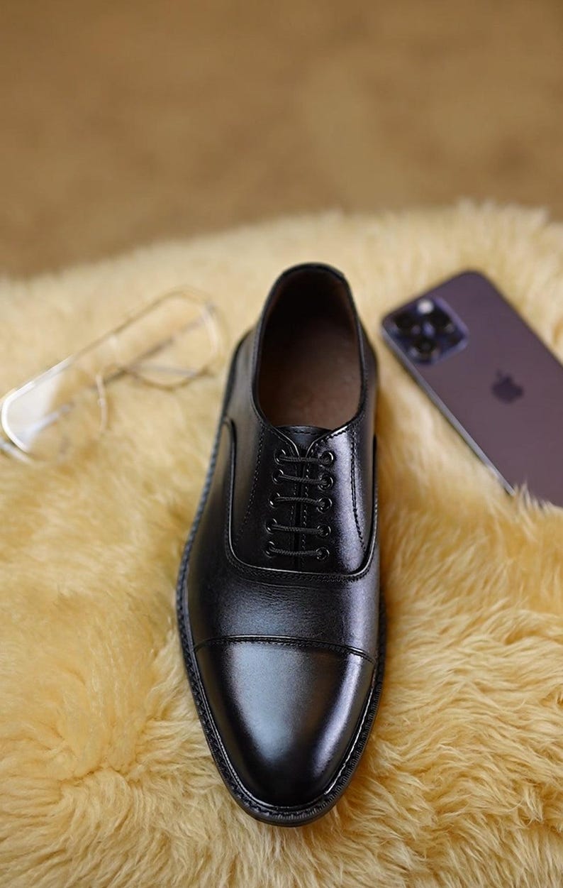 Peut inclure: Une chaussure richelieu en cuir noir avec un bout droit, dot&eacute;e d'un syst&egrave;me de la&ccedil;age ferm&eacute; avec cinq paires d'&oelig;illets et de fins lacets noirs. La chaussure pr&eacute;sente une finition lisse et polie ainsi qu'une tr&eacute;pointe cousue visible.