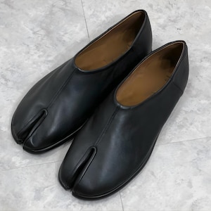 May include: A pair of black leather Tabi shoes, featuring a split-toe design. The shoes have a low profile and a smooth, polished finish. The interior is a light brown color.