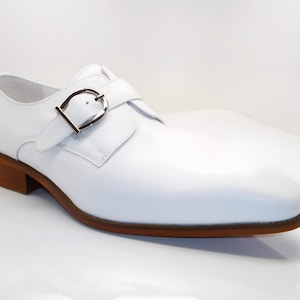 May include: A white leather shoe with a brown sole and heel. The shoe features a silver buckle closure and a classic design. The shoe is a formal style, suitable for various occasions.