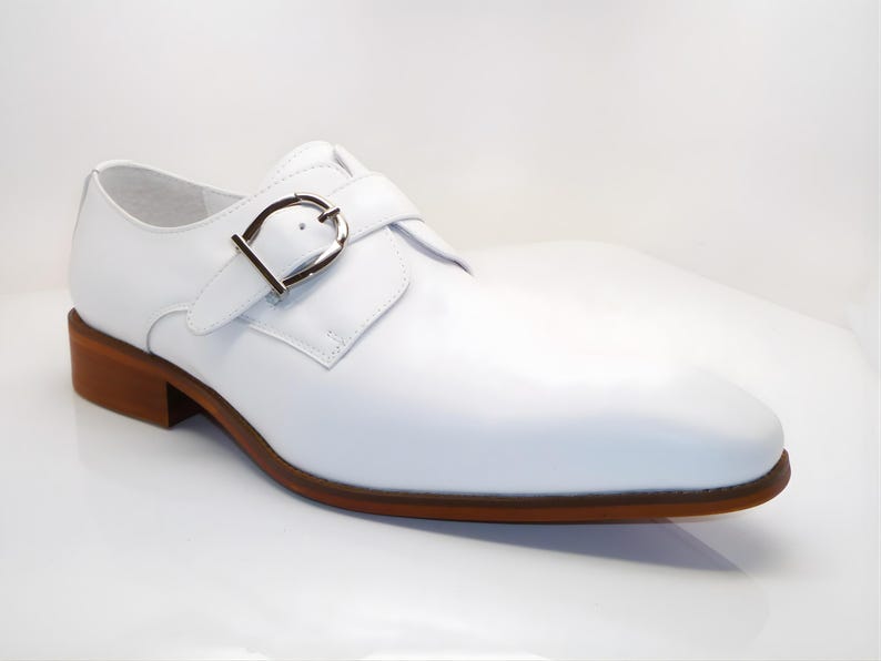May include: A white leather shoe with a brown sole and heel. The shoe features a silver buckle closure and a classic design. The shoe is a formal style, suitable for various occasions.