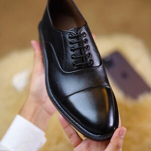 Peut inclure: Une chaussure richelieu en cuir noir avec un bout droit, un syst&egrave;me de la&ccedil;age ferm&eacute; &agrave; cinq paires d'&oelig;illets et des lacets noirs fins. La chaussure pr&eacute;sente une finition lisse et polie avec des coutures visibles et une semelle noire robuste.