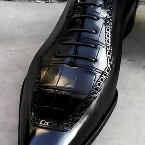 May include: A close-up of a black leather dress shoe with a glossy finish. The shoe features a textured vamp and decorative perforations along the side. The laces are black, and the shoe is on a gray surface.