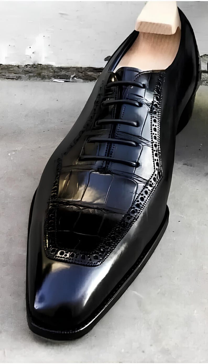 May include: A close-up of a black leather dress shoe with a glossy finish. The shoe features a textured vamp and decorative perforations along the side. The laces are black, and the shoe is on a gray surface.
