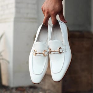 May include: A pair of white leather loafers with gold-colored metal accents. The shoes have a classic design with a rounded toe and a decorative strap across the top. The loafers are held up by a hand.