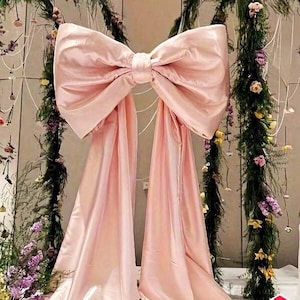 Peut inclure: Un grand nœud en satin rose clair avec de longues draperies fluides, utilisé comme élément décoratif. Le nœud est le point central, sur fond d'arrangements floraux et de verdure. L'image comprend un graphique "LIVRAISON GRATUITE".