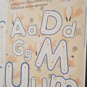 Può includere: Un quaderno per imparare l'alfabeto per bambini. La pagina presenta lettere grandi con linee tratteggiate per il tracciamento. Il testo "Learn the alphabet!" è in alto, con illustrazioni di volpi e foglie.