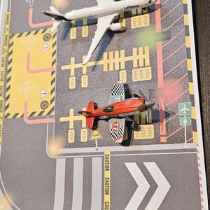 May include: A toy airplane and a red biplane sit on a printed airport runway. The white airplane has the word "Lufthansa" printed on it. The runway has caution markings and directional arrows.
