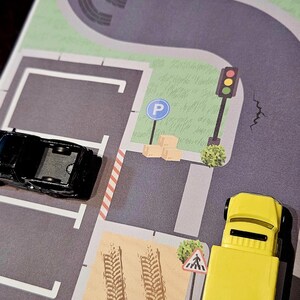 May include: A toy city scene with a road, parking area, and a pond. Includes a white and red toy car, a black toy car, and a yellow toy truck. The scene has a crosswalk and traffic light.