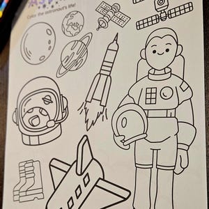 Può includere: Disegno a tratto nero e bianco di una pagina da colorare a tema astronauta. La pagina include illustrazioni di pianeti, un razzo, uno space shuttle, un satellite, un astronauta e la parola "Astronaut" in viola. È presente anche il testo "Color the astronaut's life!".