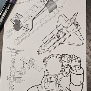 May include: A black and white line drawing of space rockets, a space station, and an astronaut. The words "Space Rockets" are at the top of the page. Two acrylic paint markers, one blue and one purple, are on the left side of the page.