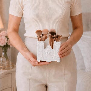 May include: A white rectangular organizer with ornate embossed floral and scrollwork details, containing a collection of makeup brushes with rose gold ferrules and brown bristles, held in a central position for storage and display purposes.