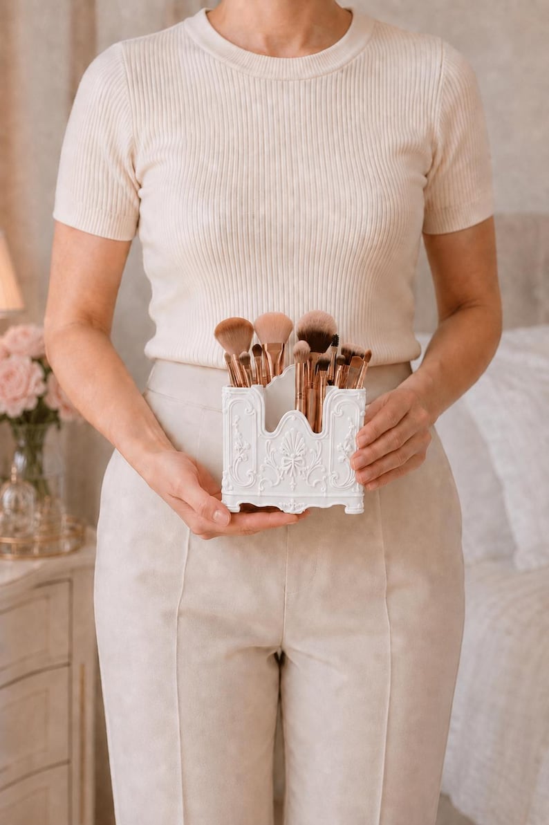 May include: A white rectangular organizer with ornate embossed floral and scrollwork details, containing a collection of makeup brushes with rose gold ferrules and brown bristles, held in a central position for storage and display purposes.