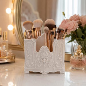 May include: A white plastic makeup brush holder with an ornate, raised relief design featuring scrollwork and a central shell motif. The container is filled with various makeup brushes that have metallic rose gold handles and soft brown bristles.
