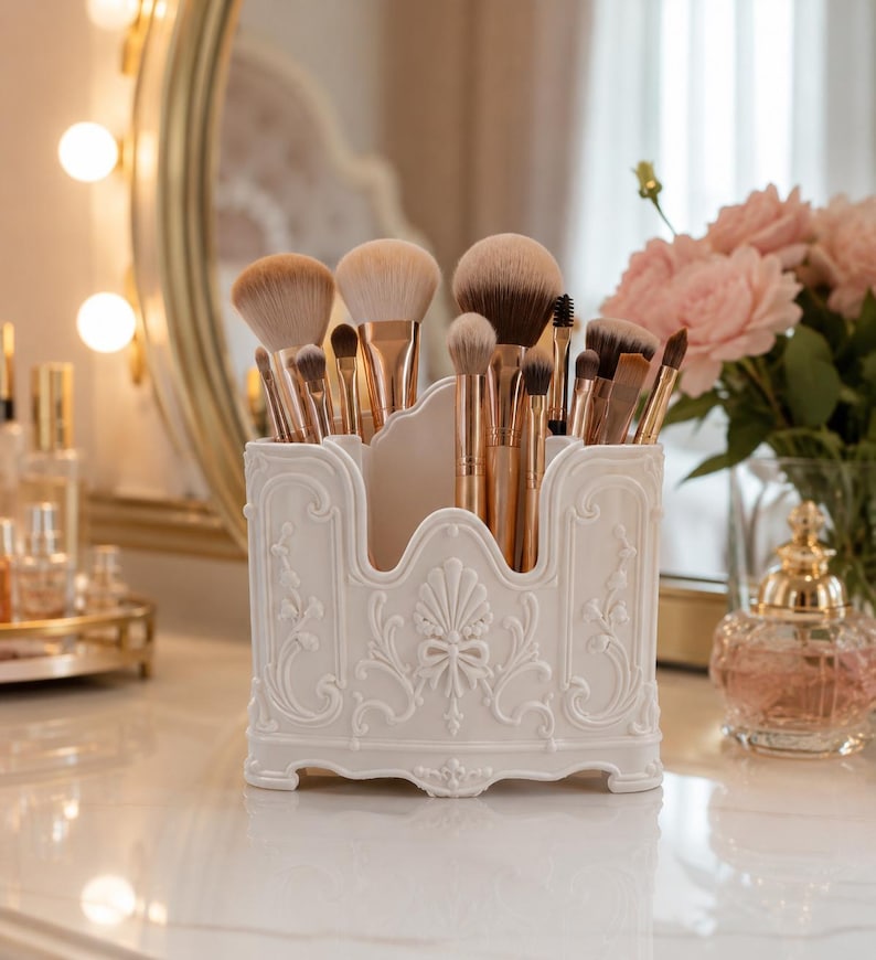 May include: A white plastic makeup brush holder with an ornate, raised relief design featuring scrollwork and a central shell motif. The container is filled with various makeup brushes that have metallic rose gold handles and soft brown bristles.