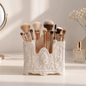 May include: A white plastic makeup brush holder featuring an ornate, raised relief design with floral and scroll motifs. The rectangular container has a scalloped top edge and holds a collection of various makeup brushes with gold-toned ferrules.